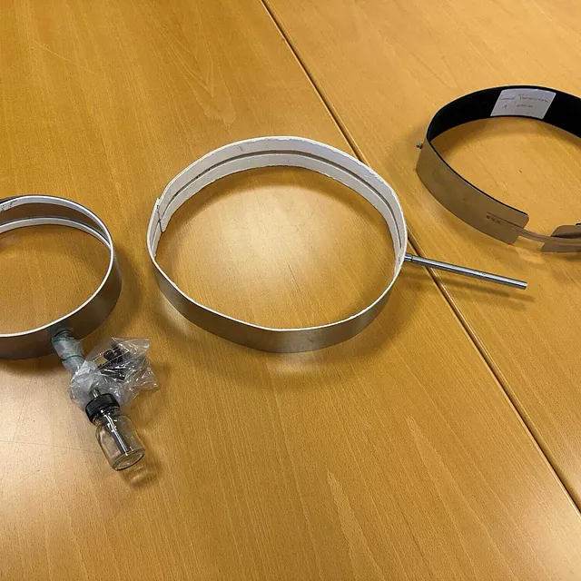 Tre metallbåndvarmere i ulike størrelser og med forskjellige tilkoblinger, lagt ut på et trebord.