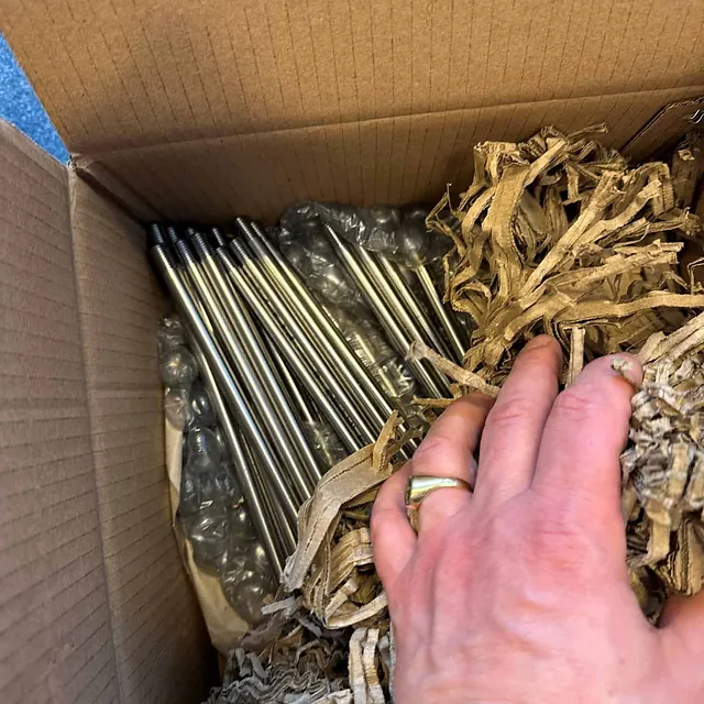 Eske fylt med stålrør med plasthetter og gjenger, delvis dekket med papirfyll og bobleplast.