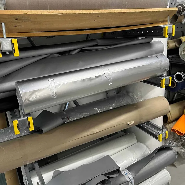 Flere ruller med teknisk tekstil og folie oppbevart på metallstativ i et lagerrom.